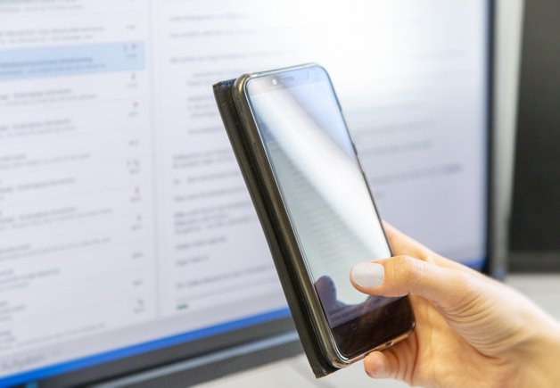 Hand hält Handy. Im Hintergrund ein Computermonitor